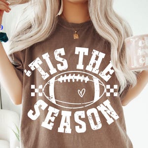 Puede incluir: Camiseta marrón con el texto blanco "'Tis the Season", con un gráfico de fútbol americano con un corazón. La camiseta es de corte holgado. La imagen también incluye logotipos de tipos de archivo SVG, PNG, EPS y DXF.