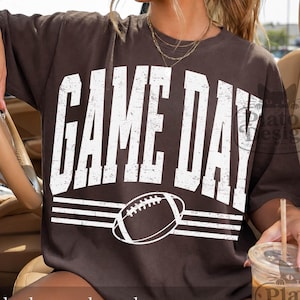 May include: A dark brown short-sleeve t-shirt featuring the white distressed text GAME DAY in a bold collegiate font. Below the text is a graphic of an American football centered between two sets of three horizontal white lines.