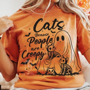 May include: An orange t-shirt featuring a black graphic print of three cats and a ghost. The text reads, "Cats Because People are Creepy." The design includes small bat silhouettes and stars, with one cat depicted as a skeleton and another as a tabby.