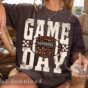 May include: A dark brown short-sleeve t-shirt featuring the text GAME DAY in large white block letters with a halftone dot pattern. A football graphic with a brown and black leopard print texture is centered between the words.