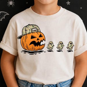 Pode incluir: Uma t-shirt de manga curta cor de creme com um gráfico de uma abóbora laranja a usar um boné de basebol camuflado, perseguindo três pequenos fantasmas com padrões de camuflagem. O design está centrado na parte frontal da camisola.