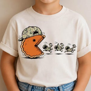 May include: A cream-colored short-sleeve t-shirt featuring a graphic of an orange pumpkin pie slice wearing a camouflage baseball cap. The pie slice is chasing three small turkeys that are running away in a line across the front of the shirt.