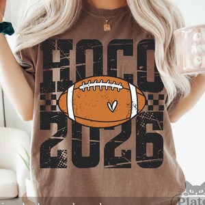 May include: A brown short-sleeve t-shirt featuring a distressed black graphic with the text HOCO 2026 and a central orange football illustration with white laces and a small heart detail. The design includes a checkered pattern element.