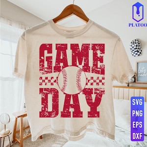 May include: A cream-coloured t-shirt featuring the words "GAME DAY" in distressed red lettering and a baseball graphic. The shirt is hanging on a wooden hanger. The design includes a baseball and a checkered pattern.