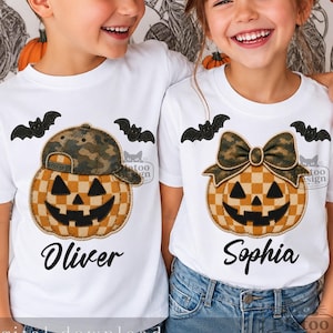 May include: Two white t-shirts featuring a checkered pumpkin graphic with a black jack-o'-lantern face and two black bats. One pumpkin wears a camouflage baseball cap above the name Oliver, and the other wears a camouflage bow above the name Sophia.