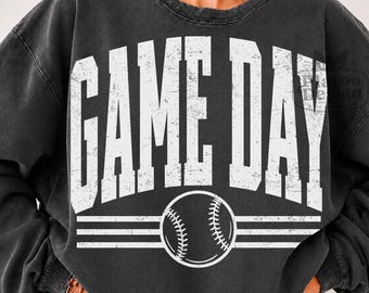 May include: A charcoal gray crewneck sweatshirt featuring the white distressed text GAME DAY in a bold athletic font, arched over a graphic of a baseball with horizontal lines extending from the sides of the ball across the chest.
