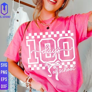 May include: A pink t-shirt with the text "100 days of school" in white. The design includes a checkered pattern and a bow. The shirt is a comfortable, casual style.