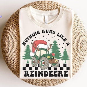 Könnte beinhalten: Cremefarbenes T-Shirt mit weihnachtlichem Motiv. Das Design zeigt einen grünen Traktor mit Weihnachtsmütze und Geweih, mit dem Text "NOTHING RUNS LIKE A REINDEERE". Enthält grüne Bäume und ein Schachbrettmuster.