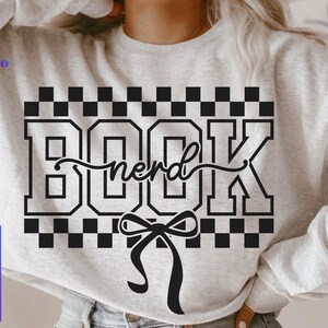 May include: A light gray sweatshirt with the word "BOOK nerd" in a black, collegiate-style font. The word "nerd" is written in a cursive font. A black bow is at the bottom of the design. The sweatshirt also has the words "SVG, PNG, EPS, DXF" in a blue and purple box.