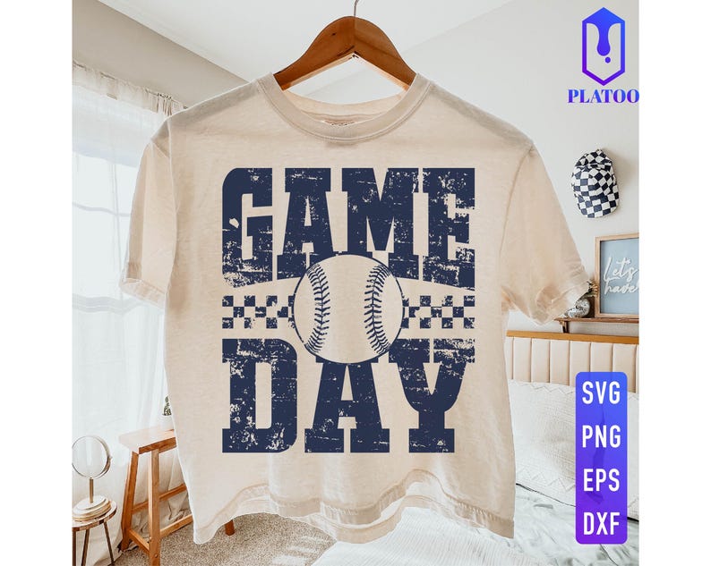 May include: A cream-coloured t-shirt with the words "GAME DAY" in distressed navy blue lettering, featuring a baseball in the centre. The shirt is hanging on a wooden hanger. The design includes a checkered pattern above and below the baseball.