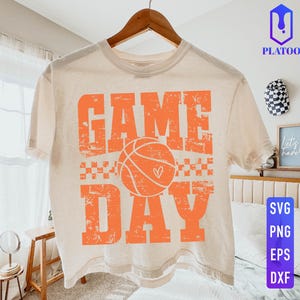 Puede incluir: Camiseta color crema con las palabras "GAME DAY" en letras naranja, con una pelota de baloncesto con un coraz&oacute;n. La camiseta est&aacute; colgada en una percha de madera.