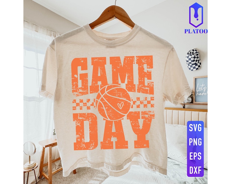Puede incluir: Camiseta color crema con las palabras "GAME DAY" en letras naranja, con una pelota de baloncesto con un coraz&oacute;n. La camiseta est&aacute; colgada en una percha de madera.