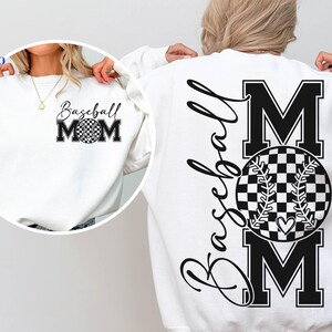 Puede incluir: Sudadera blanca con texto negro que dice "Baseball Mom" y un gráfico de béisbol. La pelota de béisbol es de cuadros negros y blancos con un pequeño corazón en el centro.