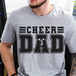 Puede incluir: Camiseta gris con texto negro que dice "Cheer Dad" con un gráfico de una animadora en el medio de la palabra "Dad".