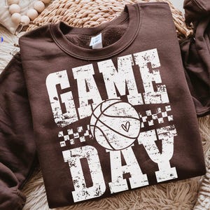 Puede incluir: Una sudadera marr&oacute;n con la frase "GAME DAY" en blanco desgastado. Un gr&aacute;fico de baloncesto con un coraz&oacute;n est&aacute; centrado entre las palabras. La sudadera es de cuello redondo.