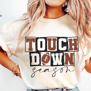 May include: Cream-colored t-shirt with the words "TOUCH DOWN season" in a brown and black design, featuring a football graphic. The shirt has short sleeves and is made of a soft material. The image also includes SVG, PNG, EPS, and DXF file type logos.