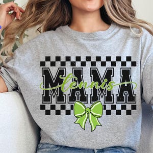 May include: A light gray t-shirt with the word "MAMA" in a bold, black, block font, with "Tennis" in a green script font. A green bow is below the text. The shirt has a checkered pattern above the word "MAMA".