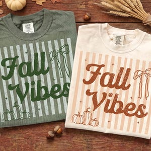 Op de afbeelding: Twee t-shirts met de tekst "Fall Vibes". Eén is saliegroen met een groen en wit ontwerp, de andere is crème met een bruin en crème ontwerp. Beide shirts hebben een strik en een pompoenafbeelding.