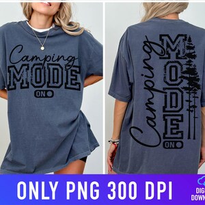 May include: A slate blue t-shirt with the words "Camping MODE ON" in black script and block letters. The back of the shirt features the same text vertically, along with a silhouette of pine trees. The shirt is a relaxed fit.
