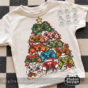 May include: A cream-coloured t-shirt featuring a graphic of a Christmas tree constructed from various video game controllers. The controllers have diverse patterns including reindeer, snowman, and Santa themes, decorated with colourful string lights.