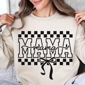 May include: Cream-colored sweatshirt with the word "MAMA" in bold, black, block letters, with a cursive "mama" overlay. The design includes a black and white checkered pattern and a black bow.