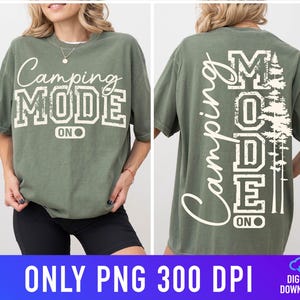 May include: An olive green t-shirt with the text "Camping Mode On" in white, in a collegiate font. The back of the shirt has the words "Camping Mode On" vertically, with a tree graphic. The shirt is a short-sleeved, casual style.