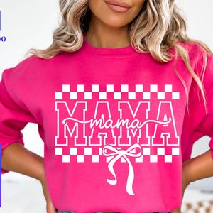 May include: A bright pink sweatshirt with the word "MAMA" in white block letters, with "mama" in cursive script underneath. A white bow is at the bottom. The sweatshirt also has a checkered pattern above the word "MAMA".