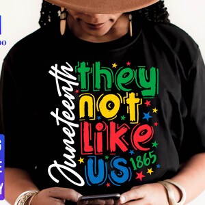 May include: A black t-shirt with the text "Juneteenth they not like us 1865" in colourful letters. The text is in a rainbow colour scheme with stars around the words.