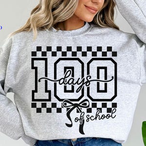 May include: A light gray sweatshirt with a black graphic that reads "100 days of school." The design includes the number 100 in a bold font, a cursive "days" and "of school" with a bow. The sweatshirt also has a checkered pattern.