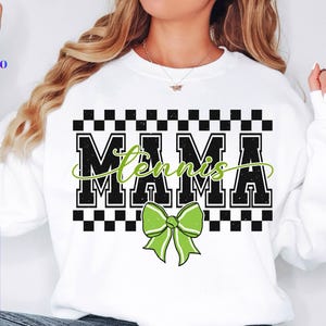 May include: White sweatshirt with the words "MAMA Tennis" in black block letters with a green cursive "Tennis" overlay. A green bow graphic is below the text. The top of the text is a black and white checkered pattern.
