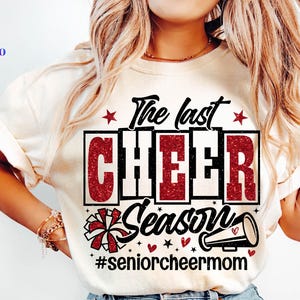 Może przedstawiać: Kremowa koszulka z napisem "The last CHEER Season" w kolorze czarnym i czerwonym. Projekt zawiera gwiazdy, megafon i pompon. Obecny jest również hashtag #seniorcheermom.