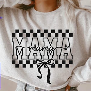 May include: A light gray sweatshirt with the word "MAMA" in large, bold, black letters, with a cursive "mama" overlay. The design includes a black and white checkered pattern and a black bow.