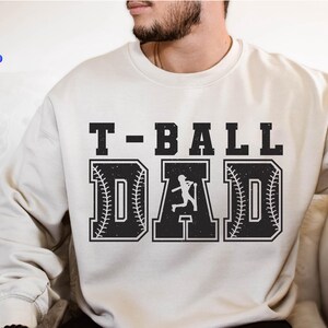 Puede incluir: Sudadera color crema con el texto "T-BALL DAD" en una fuente negra y audaz, desgastada. Las letras "D" y "A" est&aacute;n dise&ntilde;adas con costuras de b&eacute;isbol y una silueta de jugador de b&eacute;isbol. La sudadera es de estilo casual.