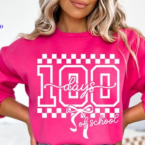 May include: A bright pink sweatshirt with the text "100 days of school" in white. The design includes the number 100, a bow, and a checkered pattern. The sweatshirt also has the words "SVG, PNG, EPS, DXF" in a vertical list.
