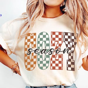 May include: Cream-colored t-shirt with the words "COZY season" in a checkered pattern. The letters are in shades of brown, green, red, and black. The shirt has short sleeves.