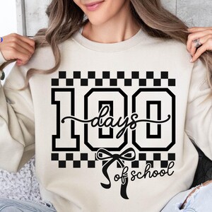 May include: A beige sweatshirt with a black graphic that reads "100 days of school." The number 100 is in a bold, blocky font, with the word "days" in a cursive script. A bow is below the word "days".