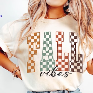May include: A cream-coloured t-shirt featuring the words "FALL vibes" in a retro checkerboard design. The letters are in shades of brown, green, and red. The shirt also has SVG, PNG, EPS, and DXF text.