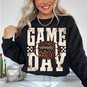May include: A black long-sleeve sweatshirt featuring the text GAME DAY in large, beige, dotted block letters. Centered between the words is a football graphic with a brown and black leopard print pattern and white laces.