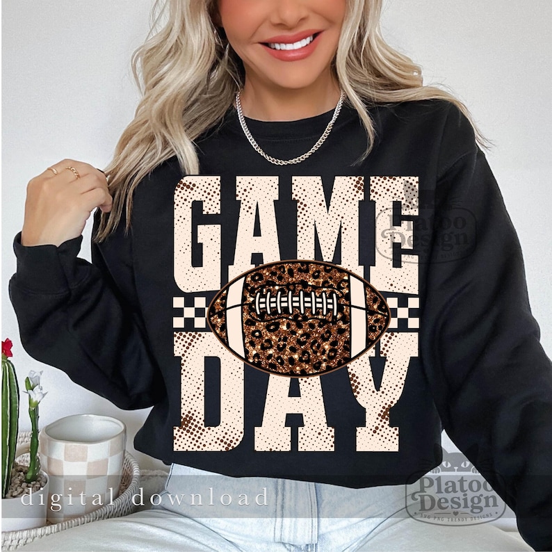 May include: A black long-sleeve sweatshirt featuring the text GAME DAY in large, beige, dotted block letters. Centered between the words is a football graphic with a brown and black leopard print pattern and white laces.