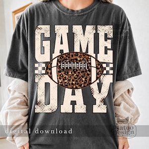 May include: A charcoal gray short-sleeve t-shirt featuring the text GAME DAY in large, cream-colored block letters with a brown halftone dot pattern. A brown and black leopard print football graphic is centered over the word GAME.