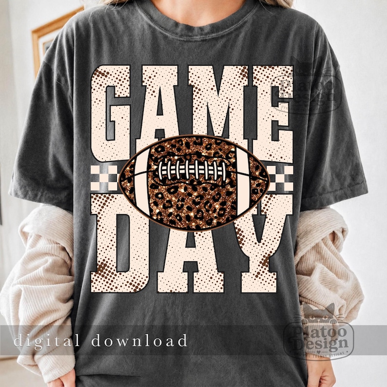 May include: A charcoal gray short-sleeve t-shirt featuring the text GAME DAY in large, cream-colored block letters with a brown halftone dot pattern. A brown and black leopard print football graphic is centered over the word GAME.