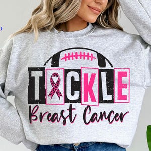 Può includere: Una felpa grigio chiaro con la scritta "TACKLE Breast Cancer" in nero e rosa. Il design include un pallone da football e un nastro rosa.