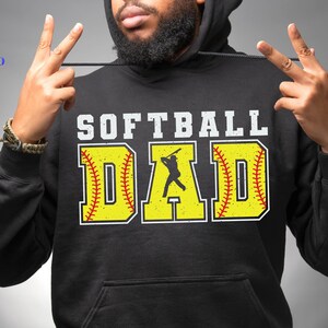 May include: Black hooded sweatshirt with the text "Softball Dad" in white letters. The text is surrounded by two yellow softball graphics with red stitching.