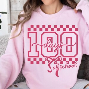 May include: A light pink sweatshirt with a graphic design in a darker shade of pink. The design features the number "100" with the words "days of school" in a script font, and a bow. The sweatshirt is a crew neck style.