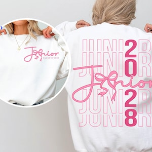 May include: A white crewneck sweatshirt featuring pink graphic text. The back displays the word JUNIOR in large outlined letters with the word Junior in cursive script and a bow graphic. The front chest shows the same cursive text and bow.