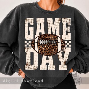 May include: A charcoal gray crewneck sweatshirt featuring a large graphic print with the text "GAME DAY" in a beige, dotted pattern. Centered between the words is a football illustration with a brown and black leopard print texture.