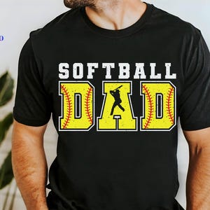 Op de afbeelding: Zwart T-shirt met de tekst "Softball Dad" in witte letters. De tekst wordt omringd door twee gele softball-afbeeldingen met rode stiksels.