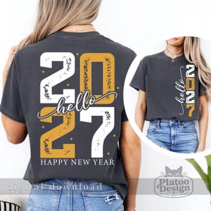 Puede incluir: Una camiseta gris carb&oacute;n con un gr&aacute;fico grande en la espalda con el texto "hello 2027" y "HAPPY NEW YEAR" en letras blancas y doradas desgastadas, con peque&ntilde;as estrellas. El &aacute;rea del pecho muestra el texto "hello 2027" verticalmente.