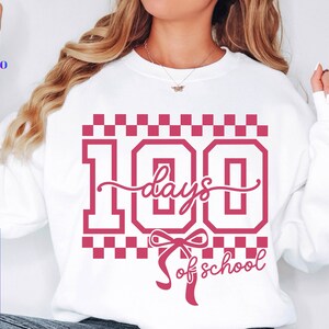 May include: White sweatshirt with a pink graphic that reads "100 days of school." The design includes the number 100 in a bold font, with the words "days" and "of school" in a script font. The design is surrounded by a checkered pattern.