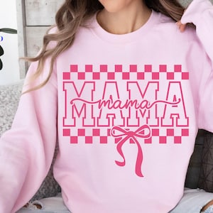 May include: A light pink sweatshirt with the word "MAMA" in a pink block letter design, with a pink bow at the bottom. The word "mama" is in a cursive font. The sweatshirt has a checkered pattern above the word "MAMA".